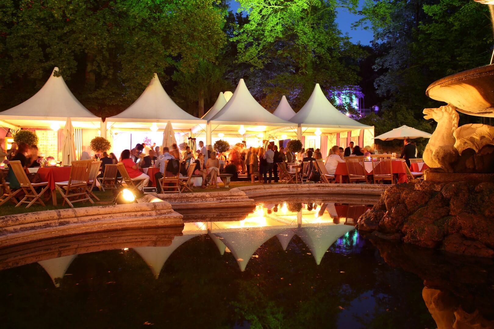 Menschen besuchen eine abendliche Veranstaltung im Freien mit Zelten, Tischen und Stühlen, die um einen reflektierenden Brunnen unter beleuchteten Bäumen aufgestellt sind. Die Szene ist durch künstliches Licht lebendig.