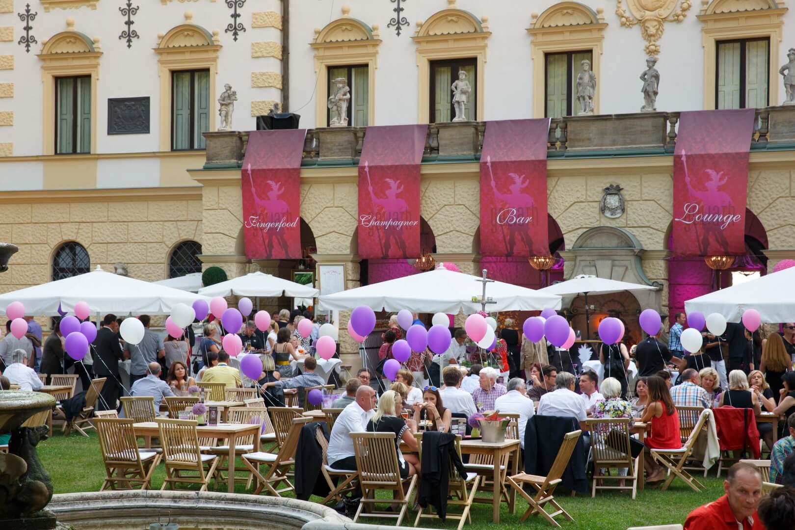 Veranstaltung im Freien mit Menschen, die an Tischen unter weißen Sonnenschirmen sitzen. Rosa und lila Luftballons und große Banner mit den Aufschriften „Lounge“, „Bar“ und „Champagner“ schmücken ein großes Gebäude im Hintergrund.