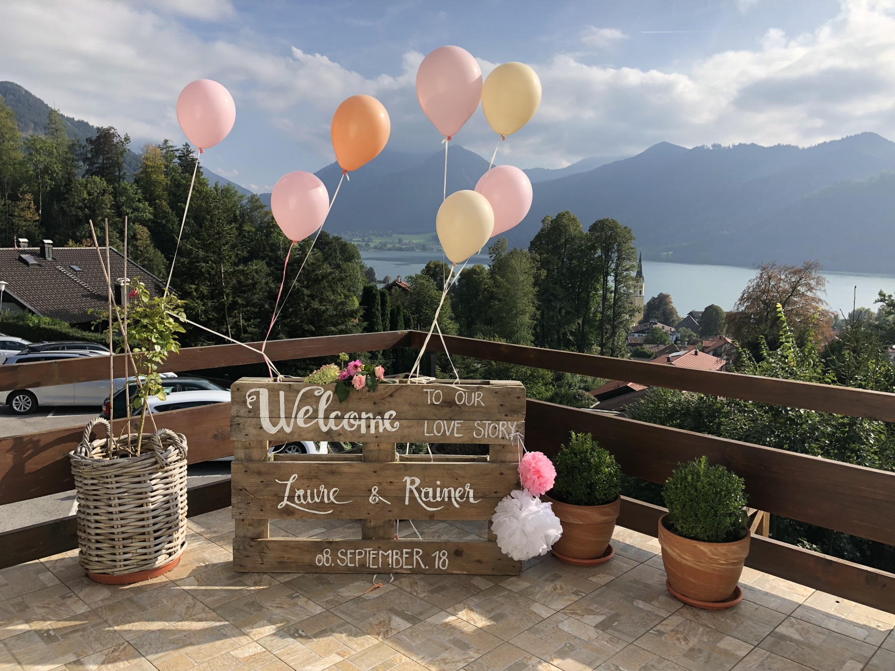 Ein Holzschild mit der Aufschrift „Willkommen zu unserer Liebesgeschichte“, Namen und Datum schmückt eine Terrasse mit Blick auf einen malerischen See und die Berge. Rosa und weiße Luftballons, Topfpflanzen und ein Weidenkorb bereichern das Dekor.