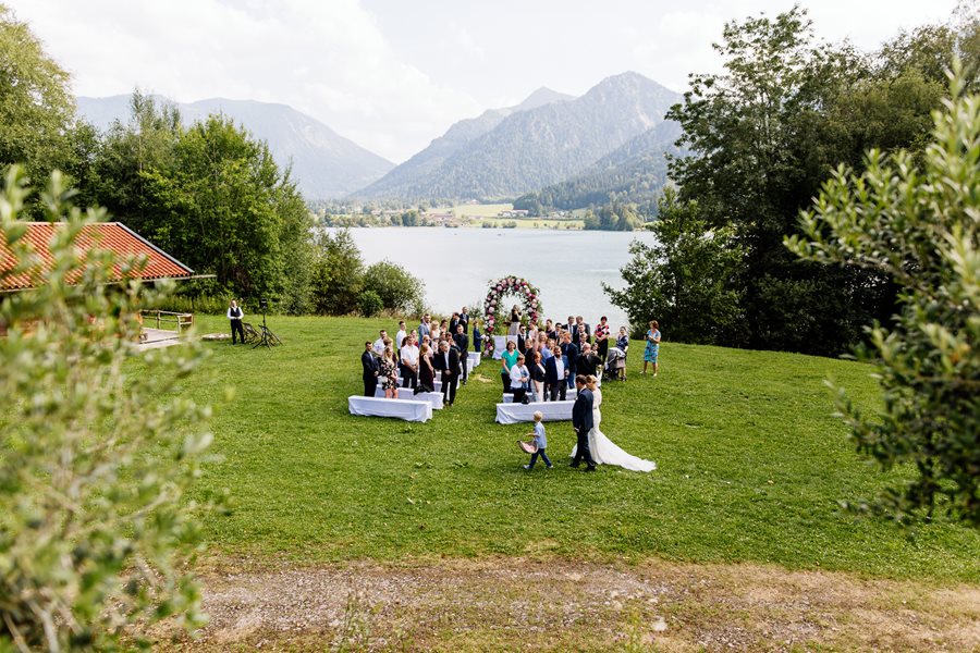 Hochzeitszeremonie im Freien auf einer Rasenfläche mit sitzenden Gästen, flankiert von Bergen und einem See im Hintergrund.