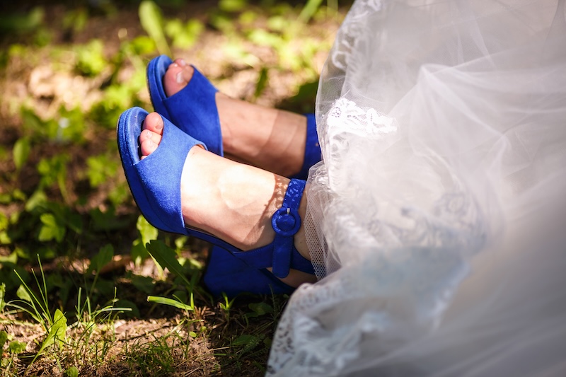 Braut trägt leuchtend blaue Sandalen mit hohen Absätzen unter einem weißen Brautkleid und steht auf Gras, durch das das Sonnenlicht fällt.